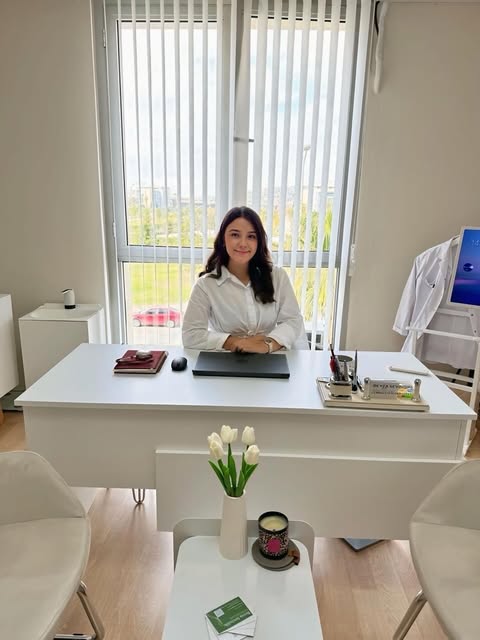 Fizyoterapist Beyza Sevindik, Mavişehir'deki kliniğinde beyaz çalışma masasının başında oturuyor; arkasında pencere ve beyaz önlüğü var.